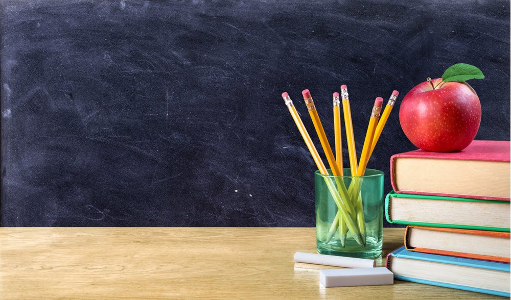 A school desk with chalk, a glass holding 6 pencils and a stack of 4 books on top. An apple is on top of the stack of books. 